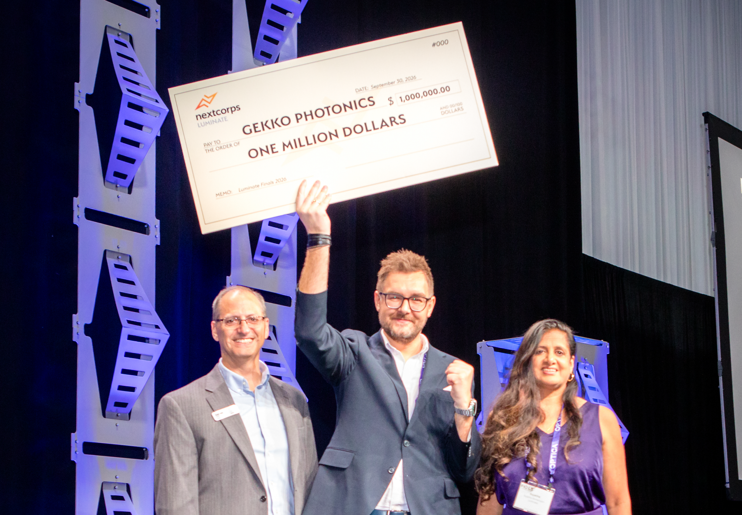 Three people stand on a stage holding a giant novelty check for $1,000,000 made out to "GEKKO PHOTONICS" from NextCorps Luminate. The man in the center holds the check aloft with one hand while raising his other fist in celebration, flanked by a man on his left and a woman on his right.