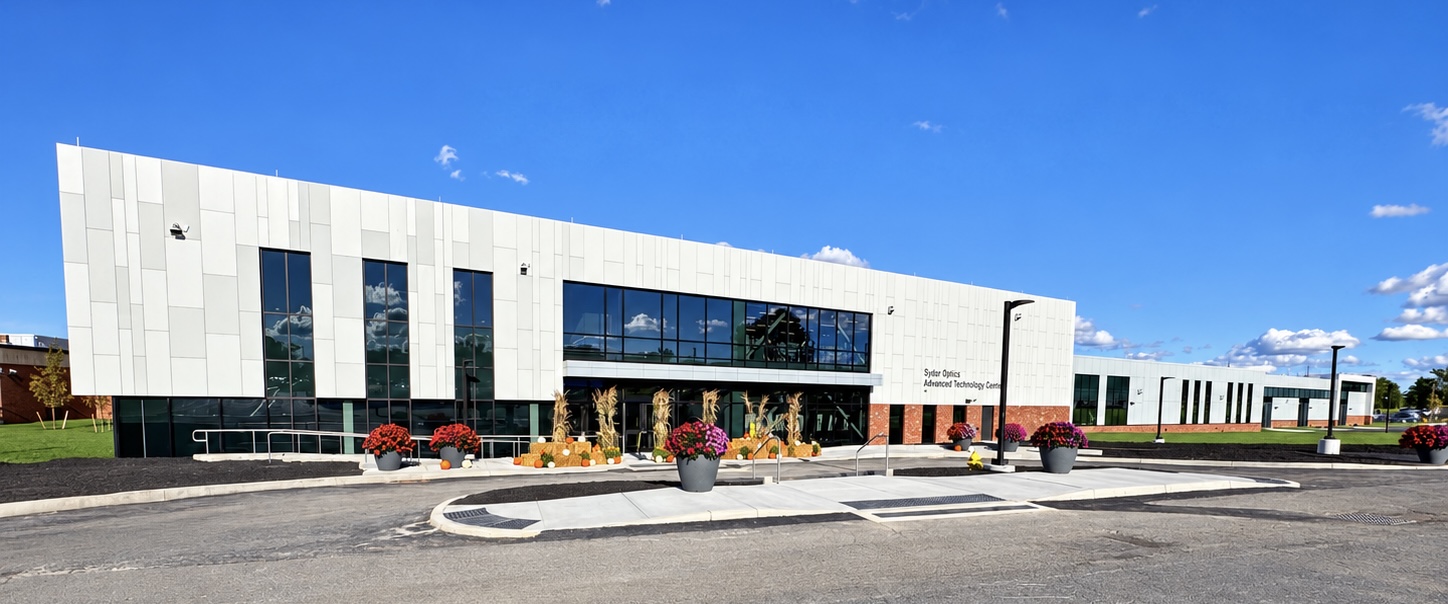A modern, light gray two-story building featuring large dark glass windows and paneling under a clear blue sky. The entrance is decorated with autumn accents, including cornstalks, pumpkins, and large planters filled with colorful flowers along the paved walkway and front apron. Signage on the brick facade reads "Sydor Optics Advanced Technology Center".